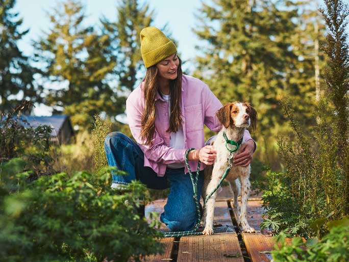 A woman in a green beanie walking her Brittany Spaniel through a foresty area